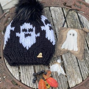 Op de afbeelding: Een zwart-witte gebreide muts met een spookontwerp en een zwarte pompon. De muts ligt op een houten tafel met een spookvormige banner van jute en een klein wit spookversiering.
