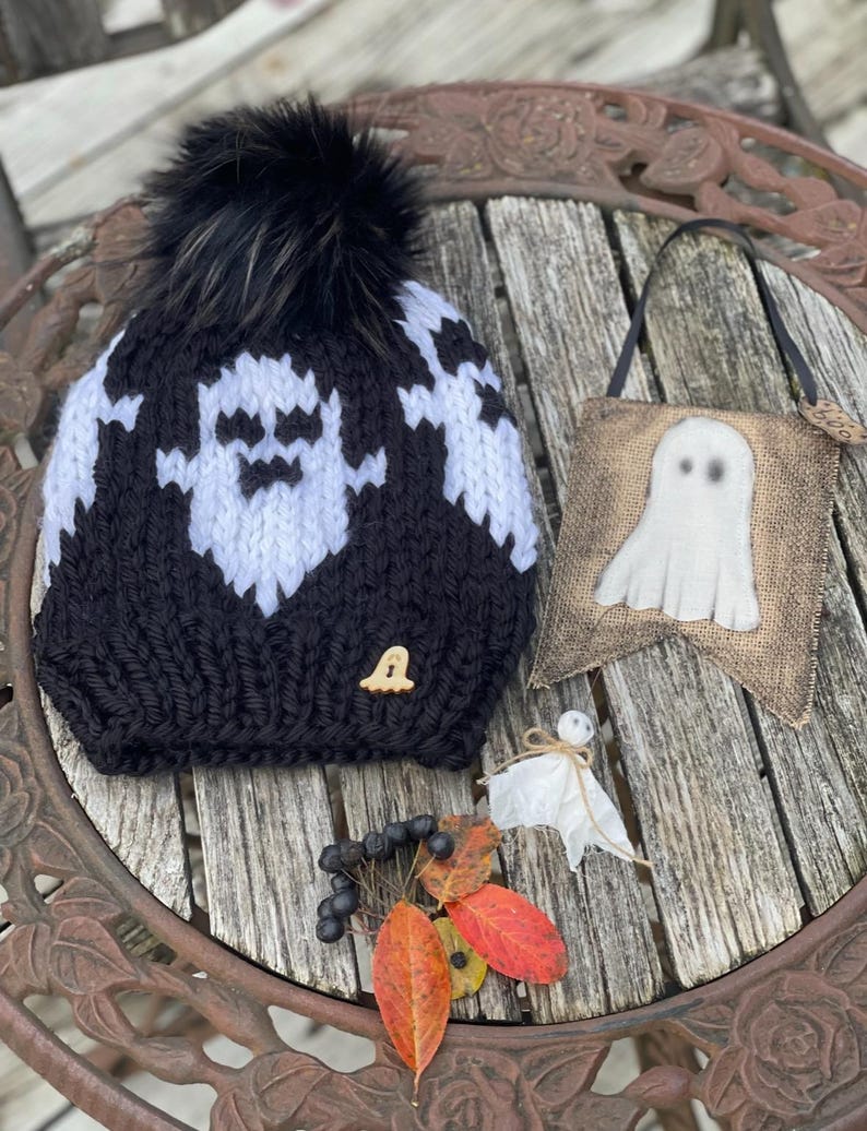 Op de afbeelding: Een zwart-witte gebreide muts met een spookontwerp en een zwarte pompon. De muts ligt op een houten tafel met een spookvormige banner van jute en een klein wit spookversiering.