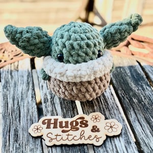 May include: A crocheted plush toy in the likeness of a character with large ears, in shades of green, beige, and brown. The toy has black eyes and a white collar. A wooden sign with the words "Hugs & Stitches" is in front of the toy.