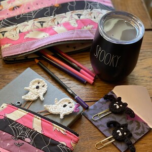 May include: A pink and black quilted zippered pouch with a Halloween theme. The pouch is open and has pencils inside. There are two crocheted ghost bookmarks and a black and white crocheted bat bookmark on a grey notebook. A black tumbler with the word "Spooky" on it is on a wooden table.