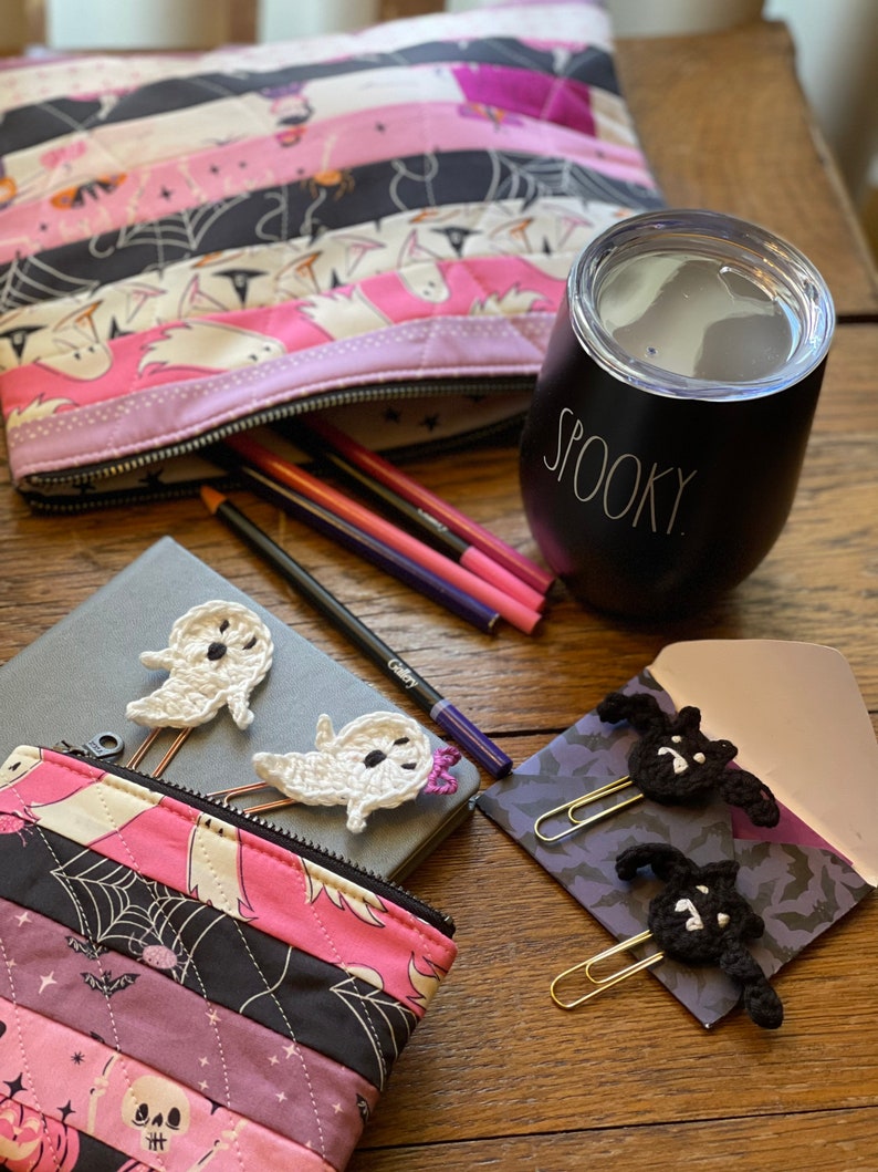 May include: A pink and black quilted zippered pouch with a Halloween theme. The pouch is open and has pencils inside. There are two crocheted ghost bookmarks and a black and white crocheted bat bookmark on a grey notebook. A black tumbler with the word "Spooky" on it is on a wooden table.