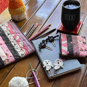 May include: A wooden table with a variety of Halloween-themed crafts, including a crocheted ghost, a crocheted candy corn, a ghost-shaped paperclip, and a black and white crocheted cat. There are also two zippered pouches with a pink and black patchwork design, a black mug with the word "Spooky" on it, and a ball of white yarn.