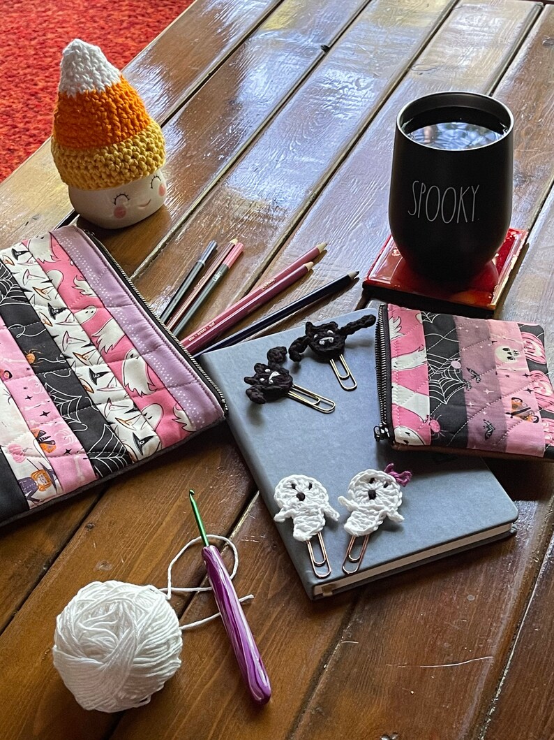 May include: A wooden table with a variety of Halloween-themed crafts, including a crocheted ghost, a crocheted candy corn, a ghost-shaped paperclip, and a black and white crocheted cat. There are also two zippered pouches with a pink and black patchwork design, a black mug with the word "Spooky" on it, and a ball of white yarn.