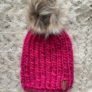 May include: A pink knitted beanie with a fluffy brown pom pom on top. The beanie has a ribbed texture and a small brown leather tag with the word "Wool & Co." on it.