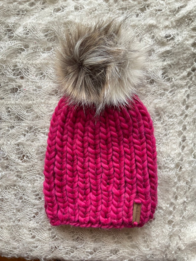 May include: A pink knitted beanie with a fluffy brown pom pom on top. The beanie has a ribbed texture and a small brown leather tag with the word "Wool & Co." on it.