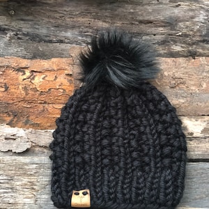 May include: Black knitted beanie with a fluffy black pom pom and a brown leather label that says "Hugs & Stitches".
