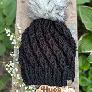 May include: A black knit beanie with a fluffy white and gray pom-pom. The hat has a textured, ribbed design and a small tag that reads "Hugs & Stitches". The hat is displayed on a wooden surface with the "Hugs & Stitches" logo and lily of the valley flowers.