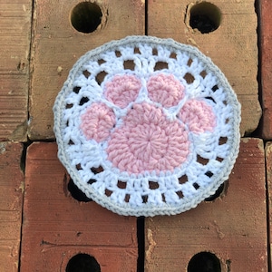 May include: A crocheted paw print coaster in pink and white with a gray border. The coaster is sitting on a brick surface.