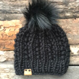 May include: Black knitted beanie with a black faux fur pom pom. The beanie has a brown leather patch with two small holes.