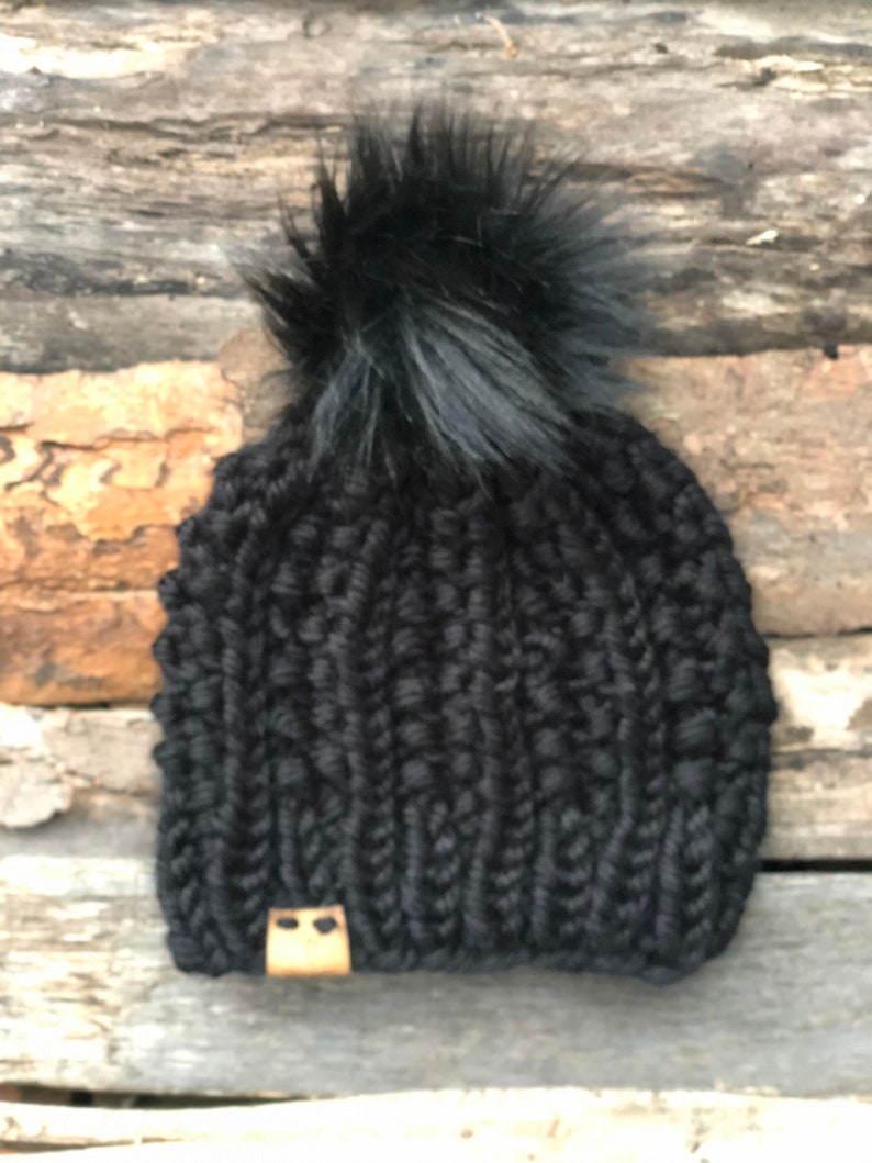 May include: Black knitted beanie with a black faux fur pom pom. The beanie has a brown leather patch with two small holes.