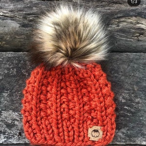 May include: A knitted beanie hat in a burnt orange color with a large fluffy pom pom in a light brown and white color. The hat has a small brown leather patch with a pumpkin design on it.