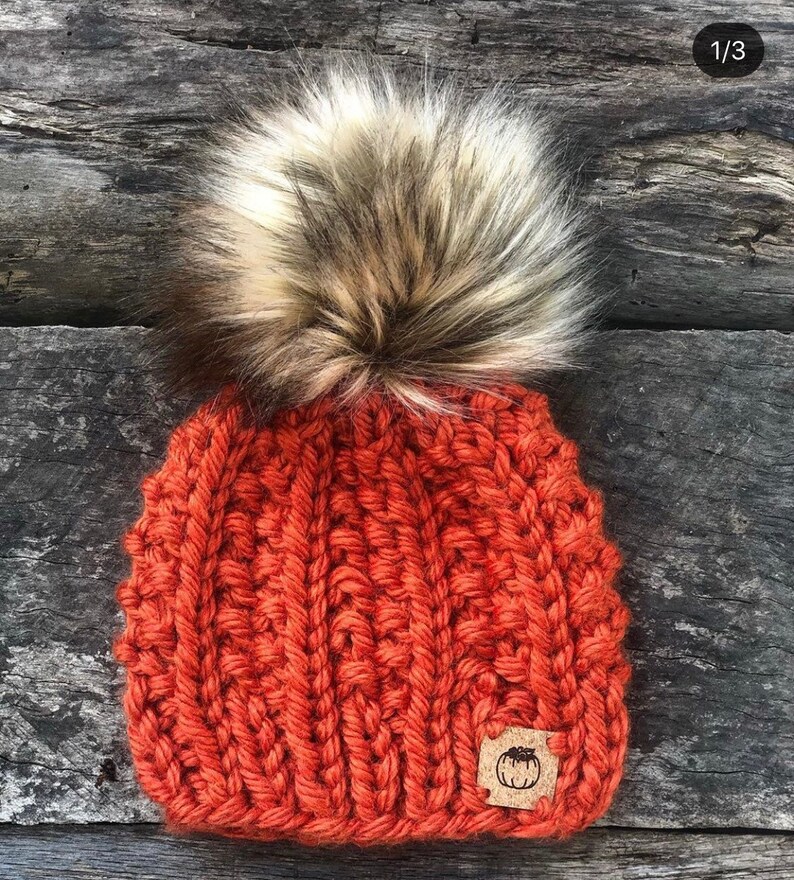 May include: A knitted beanie hat in a burnt orange color with a large fluffy pom pom in a light brown and white color. The hat has a small brown leather patch with a pumpkin design on it.