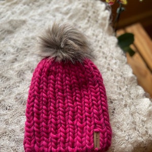 May include: A pink knitted beanie with a fluffy brown pom pom on top. The beanie has a brown leather tag with the text "Love & Co." on it.
