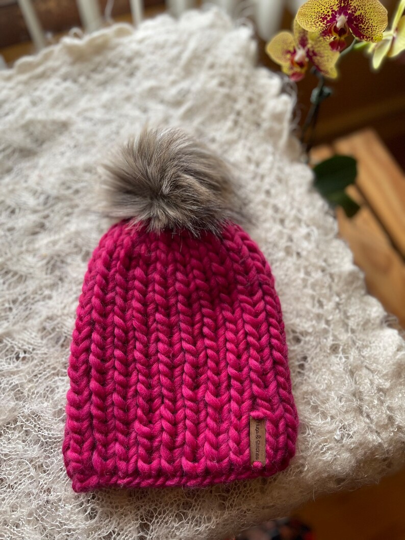 May include: A pink knitted beanie with a fluffy brown pom pom on top. The beanie has a brown leather tag with the text "Love & Co." on it.