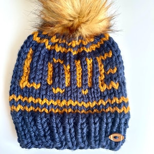 Puede incluir: Un gorro de punto azul marino con la palabra "LOVE" en hilo dorado. El gorro presenta un gran pompón de piel sintética marrón y una pequeña etiqueta de madera con forma de balón de fútbol. Un accesorio de invierno.