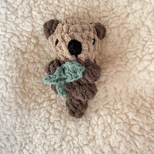 May include: A handmade crochet otter plushie in shades of brown, tan, and black, holding a light teal-colored fish. The plushie has a textured, fluffy appearance and is set against a white, textured background.