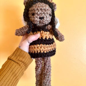 May include: A crocheted bee doll with brown yarn for the body and black yarn for the stripes. The doll has a brown face with black eyes and a small, brown nose. The doll has long, black yarn hair and white yarn wings.