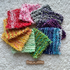 Puede incluir: Una colección de cuadrados de crochet esponjosos y coloridos en un patrón de arcoíris. Los cuadrados están dispuestos en un círculo sobre una superficie blanca y esponjosa. El texto "@hugs_art_stitches" está escrito en una etiqueta de madera en el centro inferior de la imagen.