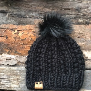 May include: Black knitted beanie with a fluffy black pom pom and a brown leather tag that says "Hues & Stitches".