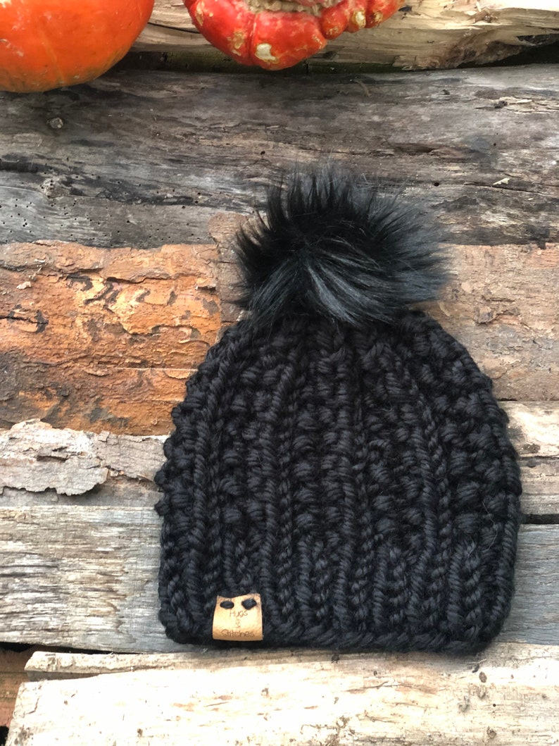 May include: Black knitted beanie with a fluffy black pom pom and a brown leather tag that says "Hues & Stitches".