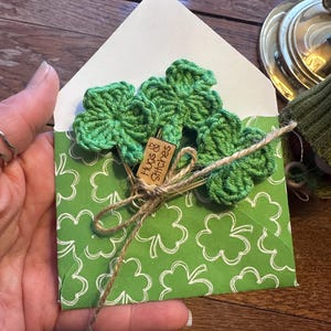 May include: A green envelope with a shamrock pattern. The envelope is tied with twine and has three green crocheted shamrock paper clips attached. The paper clips have a small wooden tag that says "Hugs & Stitches".