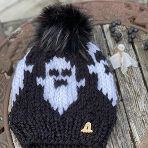 Op de afbeelding: Zwart-witte gebreide beanie met een wit spookontwerp en een zwarte nep bont pompon. De beanie heeft een kleine houten spookknop.