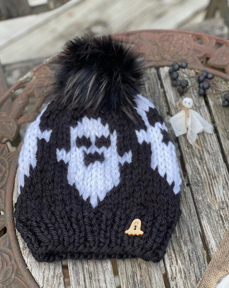 Op de afbeelding: Zwart-witte gebreide beanie met een wit spookontwerp en een zwarte nep bont pompon. De beanie heeft een kleine houten spookknop.