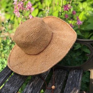 May include: A brown crocheted sun hat with a brown leather strap and a wooden toggle. The hat is sitting on a wooden bench.