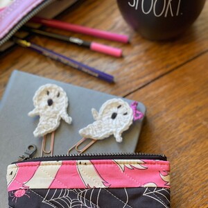 May include: A pink and black zippered pouch with a quilted design featuring bats, spiders, and spiderwebs. The pouch is partially open, revealing a grey notebook with two white crocheted ghost-shaped bookmarks.