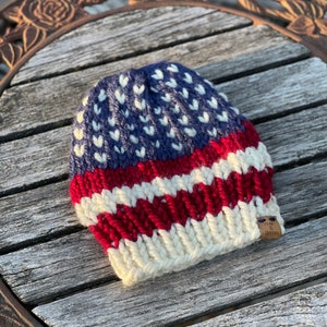 Pode incluir: Um gorro de malha com um padrão de estrelas azul e branco e listras vermelhas e brancas. O gorro tem uma pequena etiqueta de couro marrom com o texto "Hug & Stitch" nela.