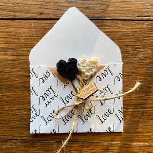 May include: White envelope with a black and white crocheted heart paperclip and a brown tag that says "Hugs & Stitches" on it. The envelope is decorated with a pattern of the word "love" written in cursive.