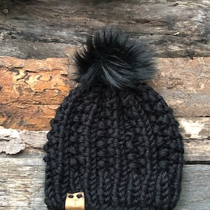 May include: Black knitted beanie with a fluffy black pom pom. The beanie has a brown leather tag with the text "The Stitches" on it.