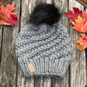 May include: A grey knitted beanie with a black faux fur pom pom. The beanie has a brown leather tag with the text "The Woolery" on it.