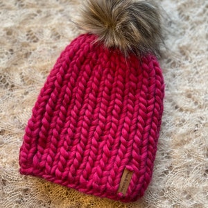 May include: A pink knitted beanie with a faux fur pom pom. The beanie has a ribbed texture and a leather tag that says "Rust & Charcoal".