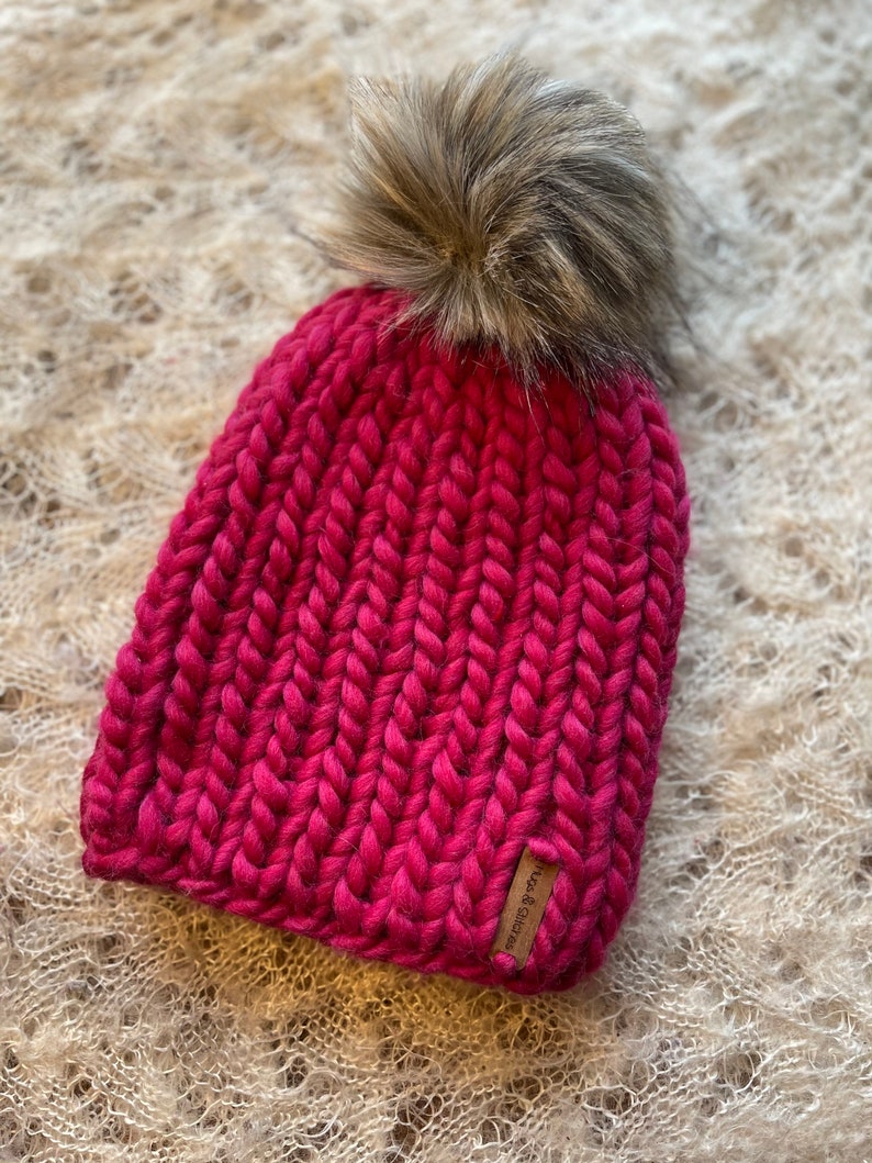 May include: A pink knitted beanie with a faux fur pom pom. The beanie has a ribbed texture and a leather tag that says "Rust & Charcoal".