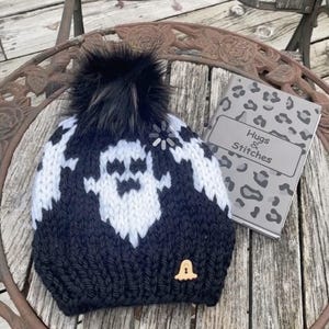 Op de afbeelding: Zwart-witte gebreide beanie met een spookontwerp en een zwarte nep bont pompon. De beanie heeft een kleine belhanger aan de zijkant. De beanie ligt op een houten oppervlak met een notitieboekje waarop "Hugs & Stitches" staat.
