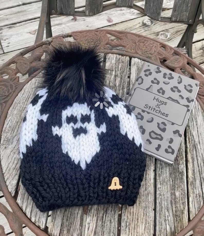 Op de afbeelding: Zwart-witte gebreide beanie met een spookontwerp en een zwarte nep bont pompon. De beanie heeft een kleine belhanger aan de zijkant. De beanie ligt op een houten oppervlak met een notitieboekje waarop "Hugs & Stitches" staat.