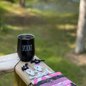 May include: A black travel mug with the word "SPOOKY" printed on it, a pink and black fabric pouch with a zipper, and several crocheted ghost and bat ornaments.