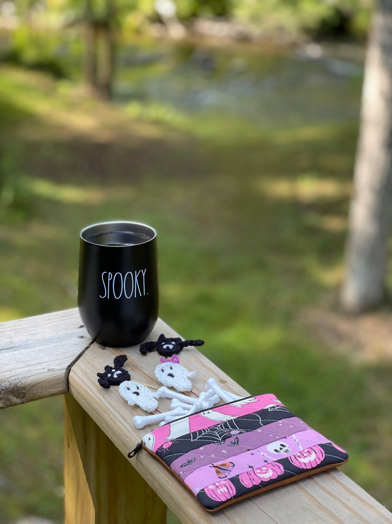 May include: A black travel mug with the word "SPOOKY" printed on it, a pink and black fabric pouch with a zipper, and several crocheted ghost and bat ornaments.