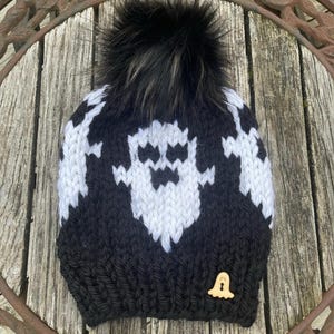 May include: Black and white knit beanie with a ghost pattern and a black faux fur pom pom. The beanie has a small ghost button on the side.