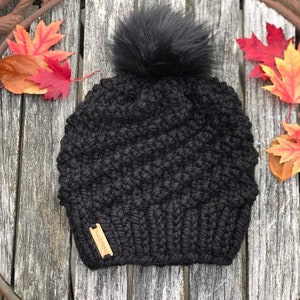 May include: Black knitted beanie with a fluffy black pom pom and a brown leather tag that says "Handmade".