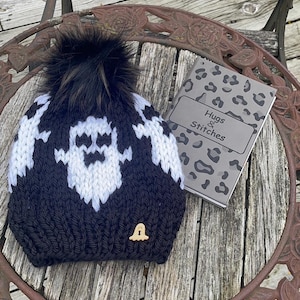 Op de afbeelding: Zwarte en witte gebreide spookmuts met een zwarte nep bont pompon. De muts heeft een kleine houten spookknop aan de zijkant. De muts ligt op een houten tafel met een notitieboekje waarop "Hugs & Stitches" staat.