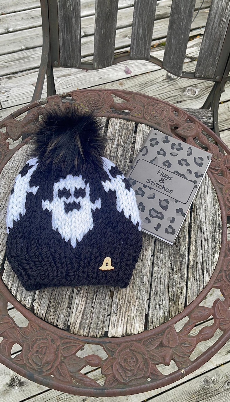Op de afbeelding: Zwarte en witte gebreide spookmuts met een zwarte nep bont pompon. De muts heeft een kleine houten spookknop aan de zijkant. De muts ligt op een houten tafel met een notitieboekje waarop "Hugs & Stitches" staat.