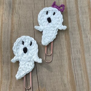 May include: Two white crocheted ghost-shaped paper clips with black eyes and mouths. One ghost has a purple bow on its head. Both are attached to rose gold paper clips.