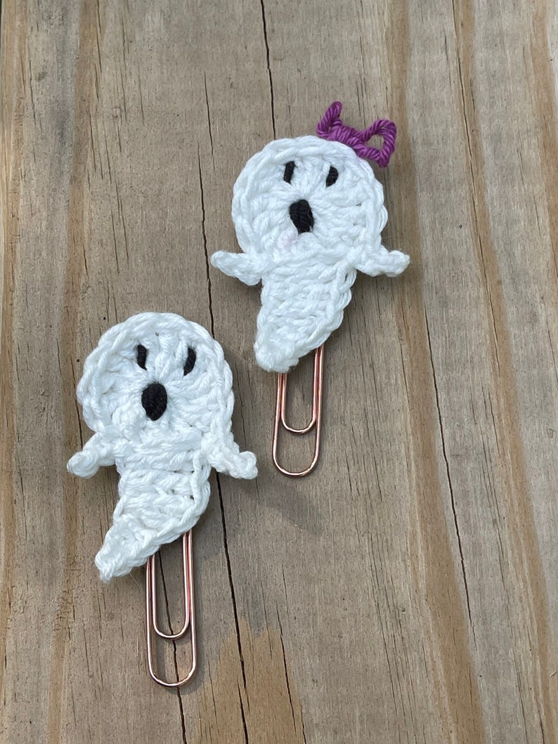 May include: Two white crocheted ghost-shaped paper clips with black eyes and mouths. One ghost has a purple bow on its head. Both are attached to rose gold paper clips.
