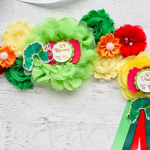 May include: A colourful baby shower sash with a green caterpillar and the text "Mommy To Be" and "Daddy To Be" on red and green circles. The sash is decorated with yellow, orange, red, and green fabric flowers.