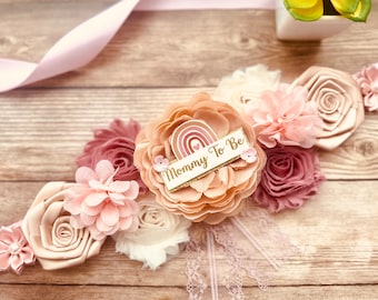 Könnte beinhalten: Ein rosa- und pfirsichfarbener Blumen-Sash mit einem Regenbogen und dem Text "Mommy To Be" in goldenen Glitzer. Der Sash ist mit Spitze und Perlen verziert.