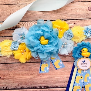 May include: A blue and yellow baby shower sash with a large blue flower and yellow flowers. The sash has a yellow ribbon with a blue circle that says "Daddy To Be" and a rubber duck on it.
