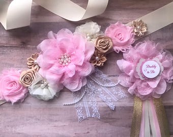 May include: A pink and gold sash with a white lace trim and a pink and white flower arrangement. The sash has a small pink and white flower with the text "Daddy to Be" in the center.