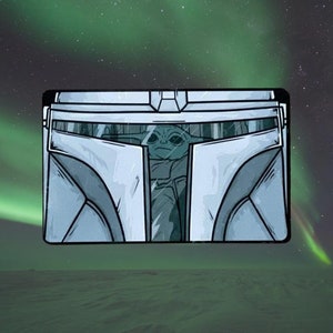 May include: Rectangular illustration featuring a stylized depiction of The Mandalorian's helmet with Grogu inside. The helmet is primarily light blue and white, with Grogu in the center. The background shows a green and purple aurora borealis.